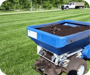 A topdressing machine filled with soil parked on a striped lawn, with work gloves resting on top.